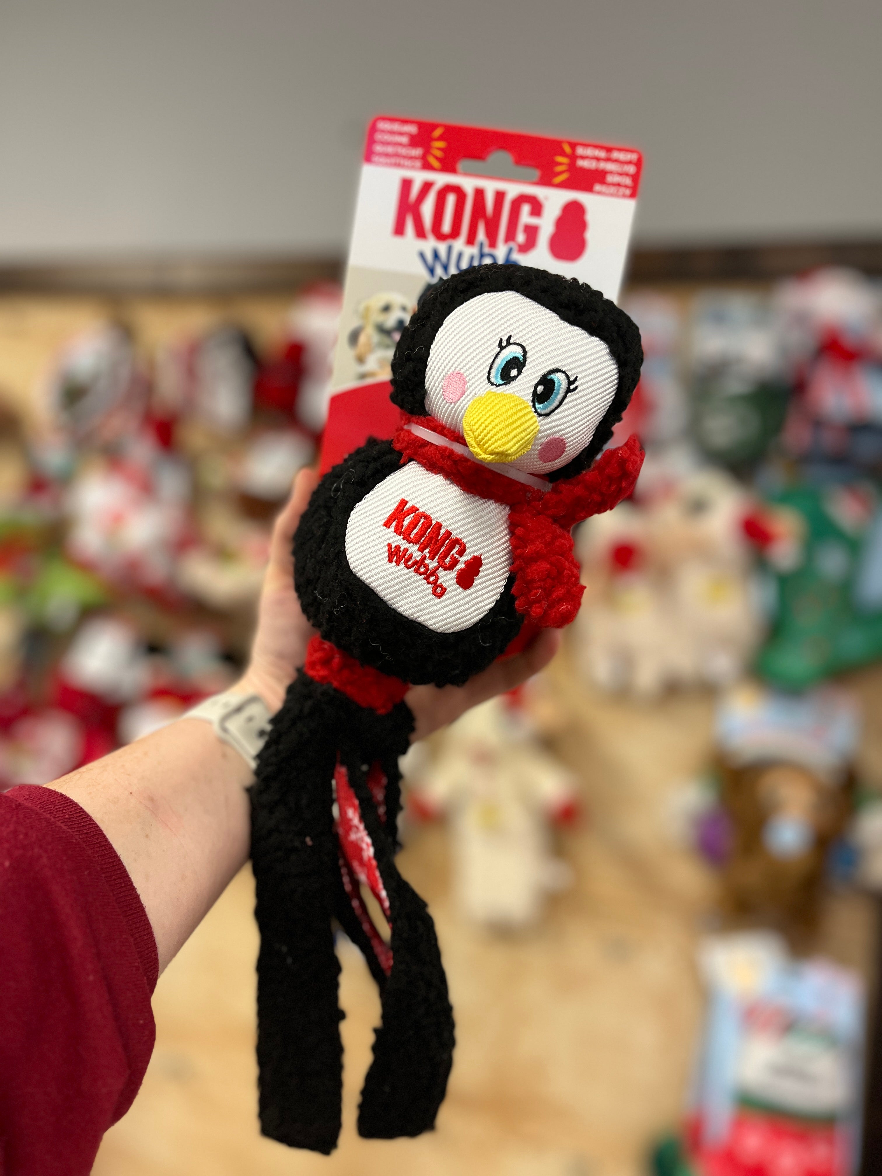 Kong Holiday Wubba toys featuring Christmas-themed characters (penguin) with soft plush, long tails for tugging, and a festive plaid design for interactive play and cozy snuggles.